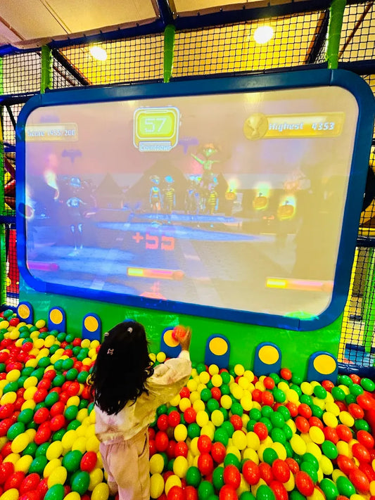 A young child plays in a ball pit in front of a large interactive screen showing a game.
