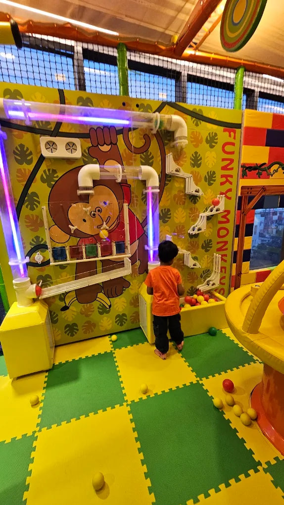 A young child plays with balls at a colorful indoor playground with a monkey-themed wall.