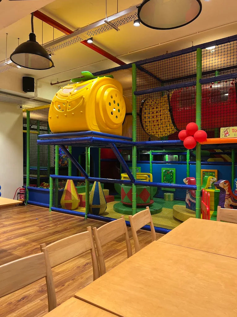 A bright indoor playground with a yellow helicopter structure and colorful play equipment. Wooden tables and chairs are in the foreground.