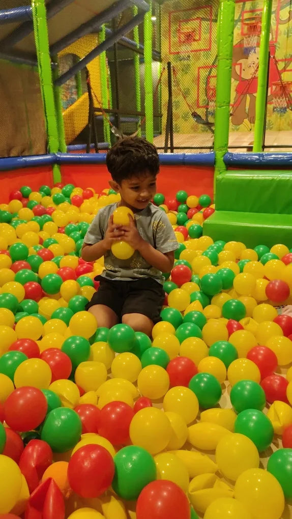 A young boy sits in a ball pit, holding a yellow ball. He is surrounded by colorful balls in a play area.