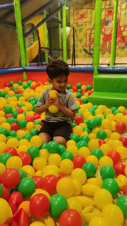A young boy sits in a ball pit, holding a yellow ball. He is surrounded by colorful balls in a play area.