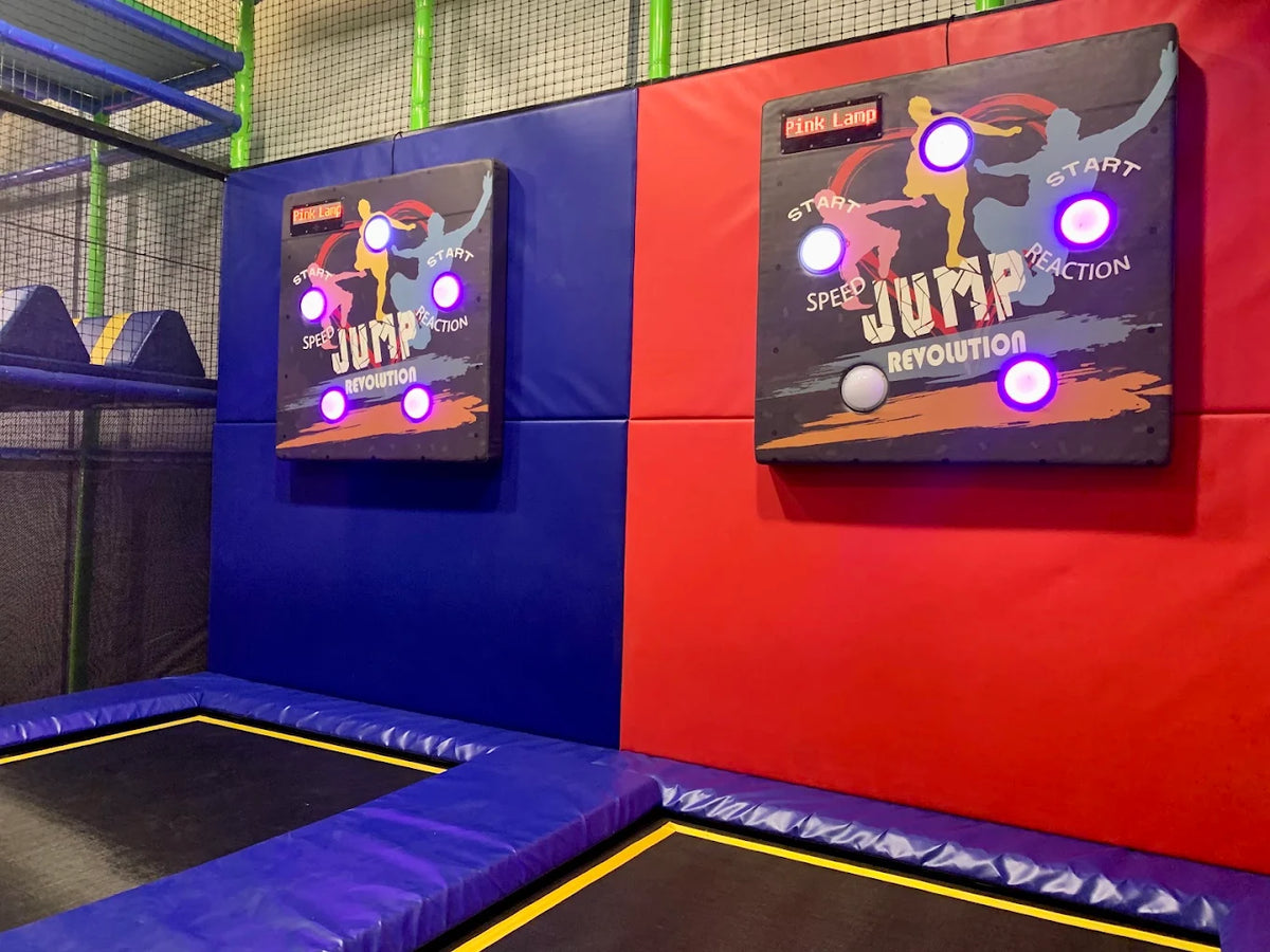 Two "Jump Revolution" game boards with illuminated buttons are mounted on a padded wall above trampolines.