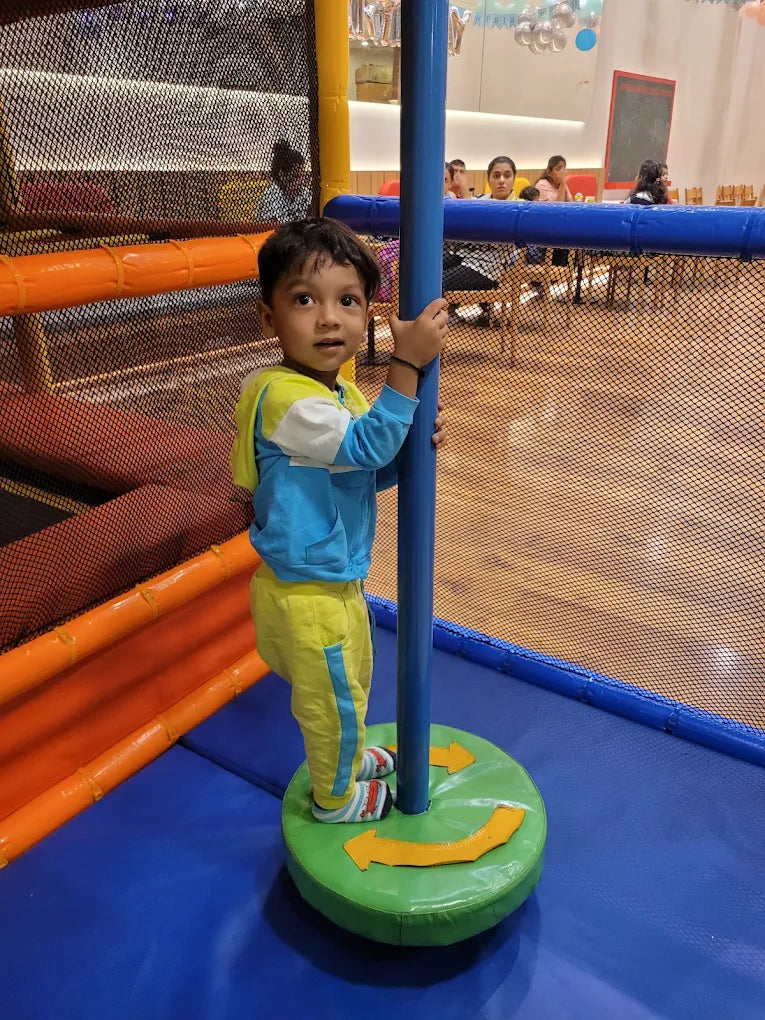 A young boy stands on a spinning platform in a colorful indoor playground. He holds onto a blue pole, looking towards the camera.