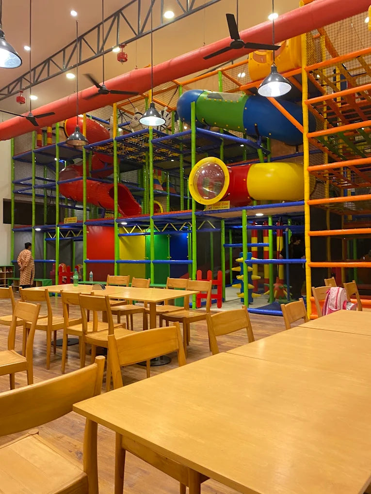 A colorful indoor playground with slides and tunnels, surrounded by wooden tables and chairs.