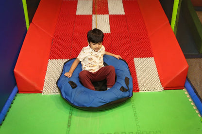 A young boy sits in a blue tube at the top of a slide in a play area. He is ready to slide down the red and white checkered surface.
