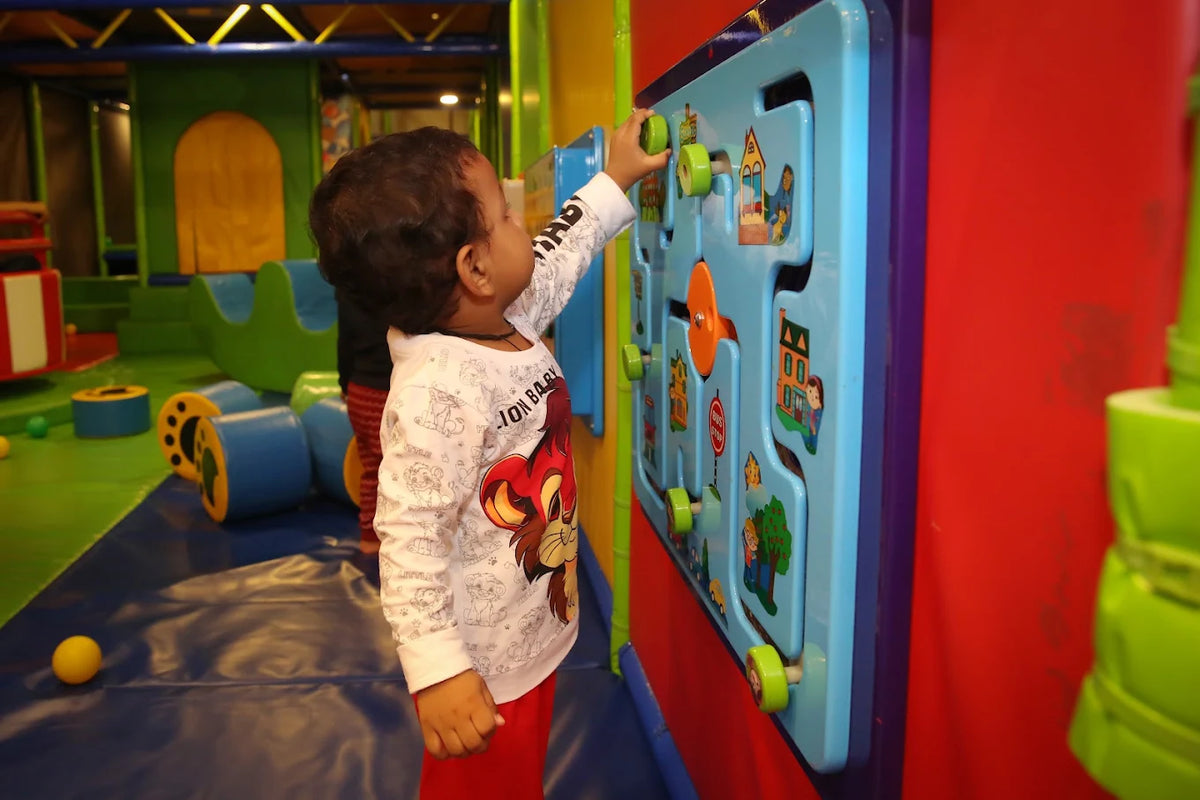 A young child plays with a colorful activity board in a brightly lit indoor playground.