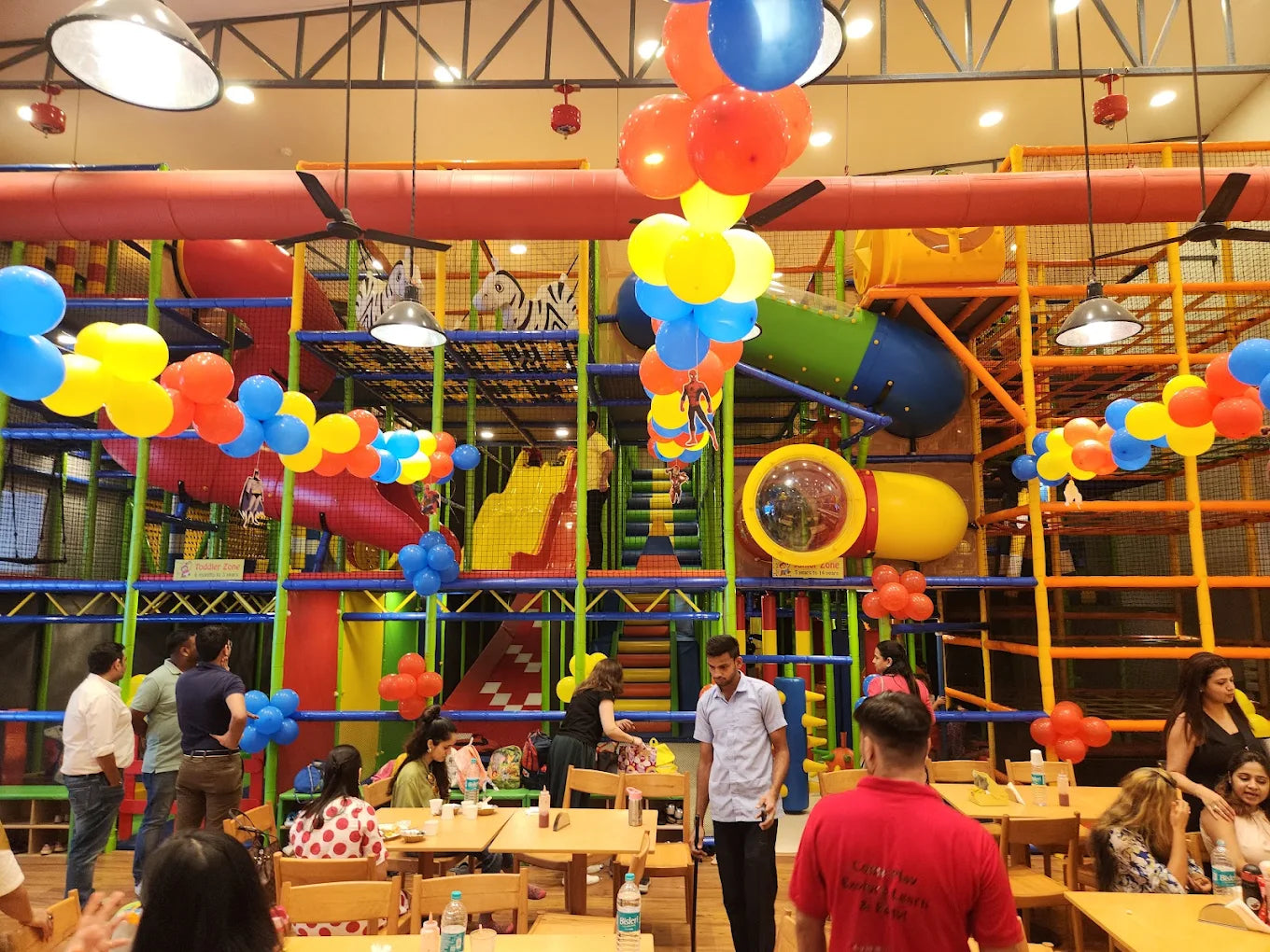 A colorful indoor playground with children and adults enjoying the space, decorated with balloons.
