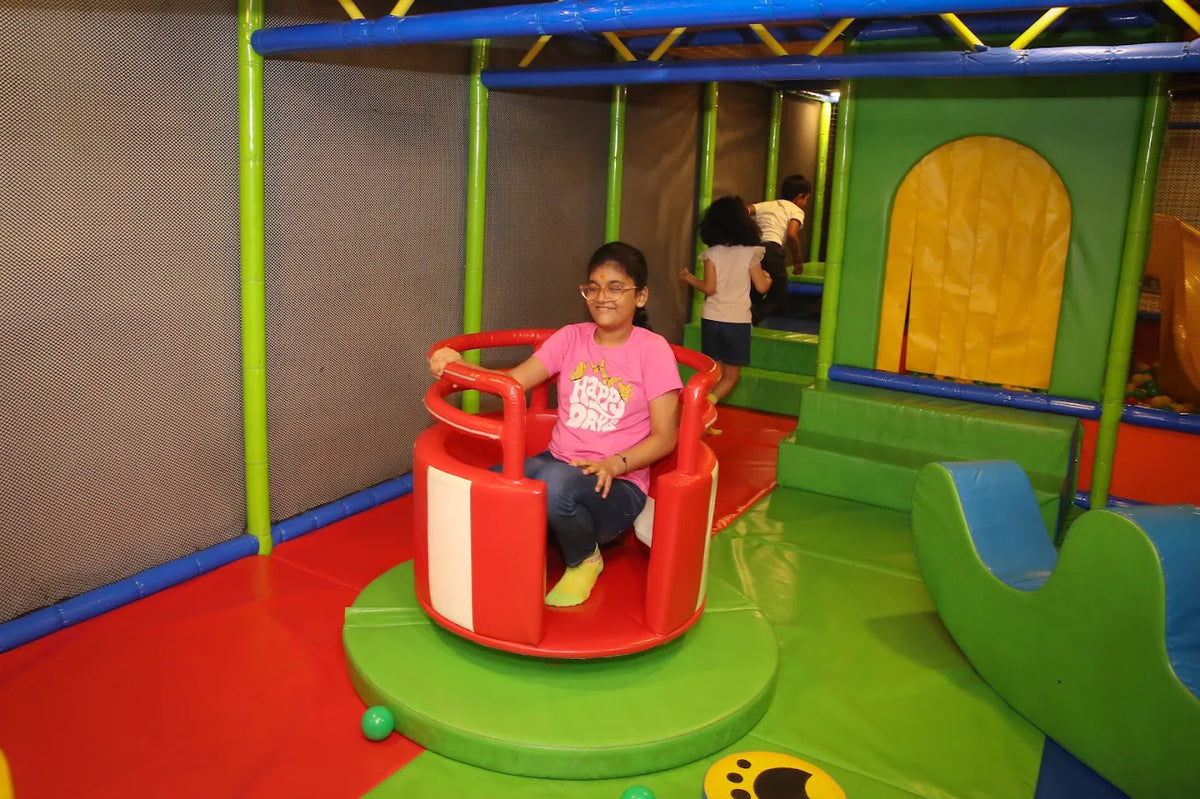 A young girl rides a red and white spinning toy in a colorful indoor playground. Other children play in the background.
