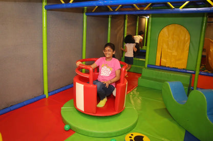 A young girl rides a red and white spinning toy in a colorful indoor playground. Other children play in the background.