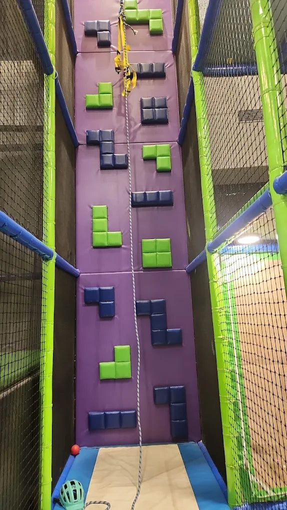 A child is climbing a colorful Tetris-themed climbing wall with a safety harness and rope.