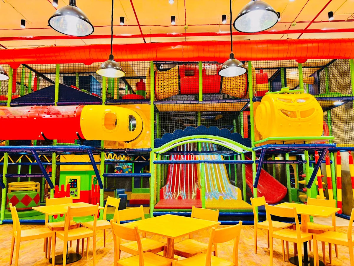 A colorful indoor playground with slides and tunnels, featuring yellow tables and chairs for children.
