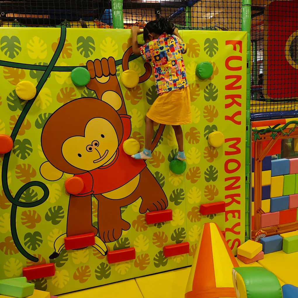 A young child climbs a colorful wall with a monkey graphic at an indoor playground.