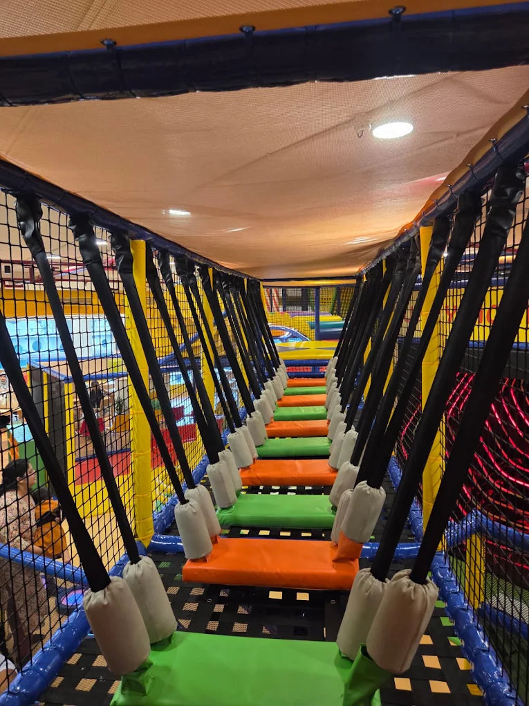 A colorful indoor playground with a suspended bridge made of orange and green steps, with children playing in the background.