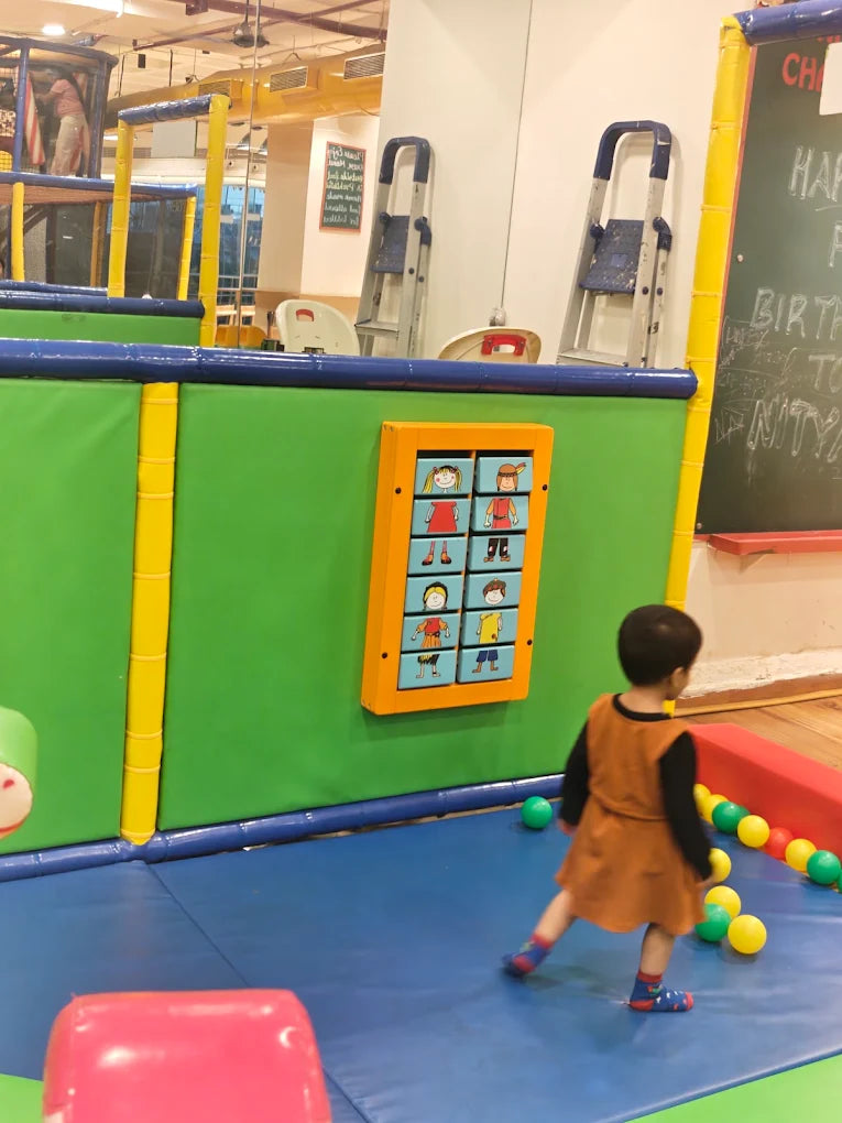 A young child plays in a colorful indoor playground with a ball pit and interactive wall game.