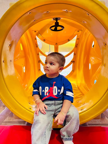 A young child sits inside a yellow tunnel slide, wearing a blue shirt with a red logo.