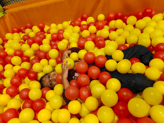 A young child smiles while lying in a ball pit filled with red and yellow balls.
