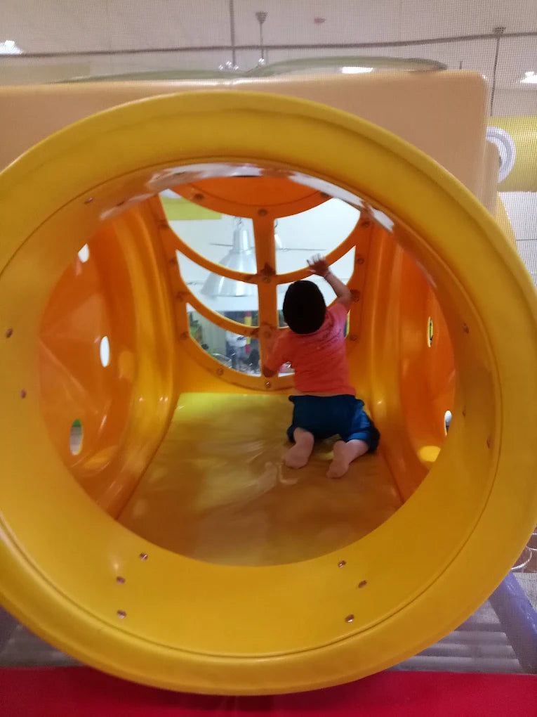 A young child crawls through a bright yellow tunnel slide in a play area.