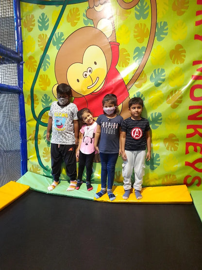 Four young children stand on a trampoline in front of a monkey-themed backdrop.