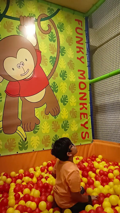 A young child sits in a ball pit, looking up at a mural of a monkey.
