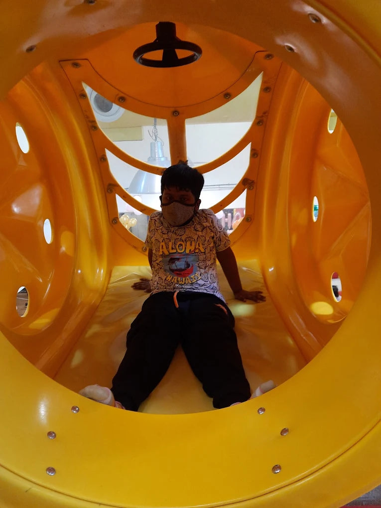 A young boy sits inside a bright yellow tunnel slide, wearing a mask and a patterned shirt.