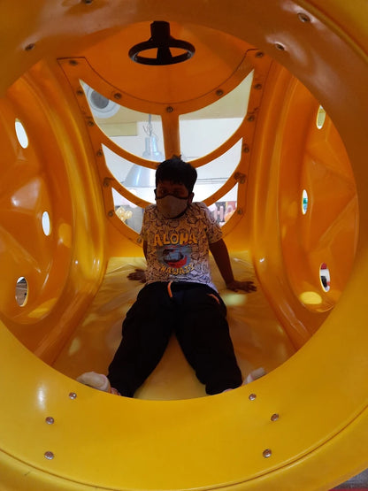 A young boy sits inside a bright yellow tunnel slide, wearing a mask and a patterned shirt.