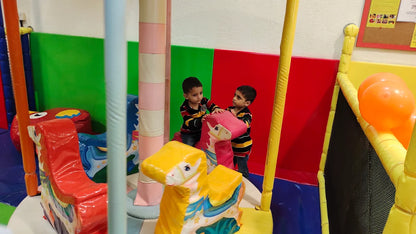Two young boys play on colorful rocking horses in a soft play area.