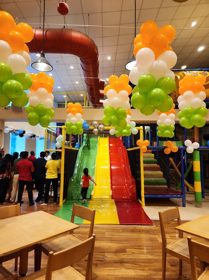 A child slides down a colorful play structure while adults watch, with balloons decorating the indoor play area.