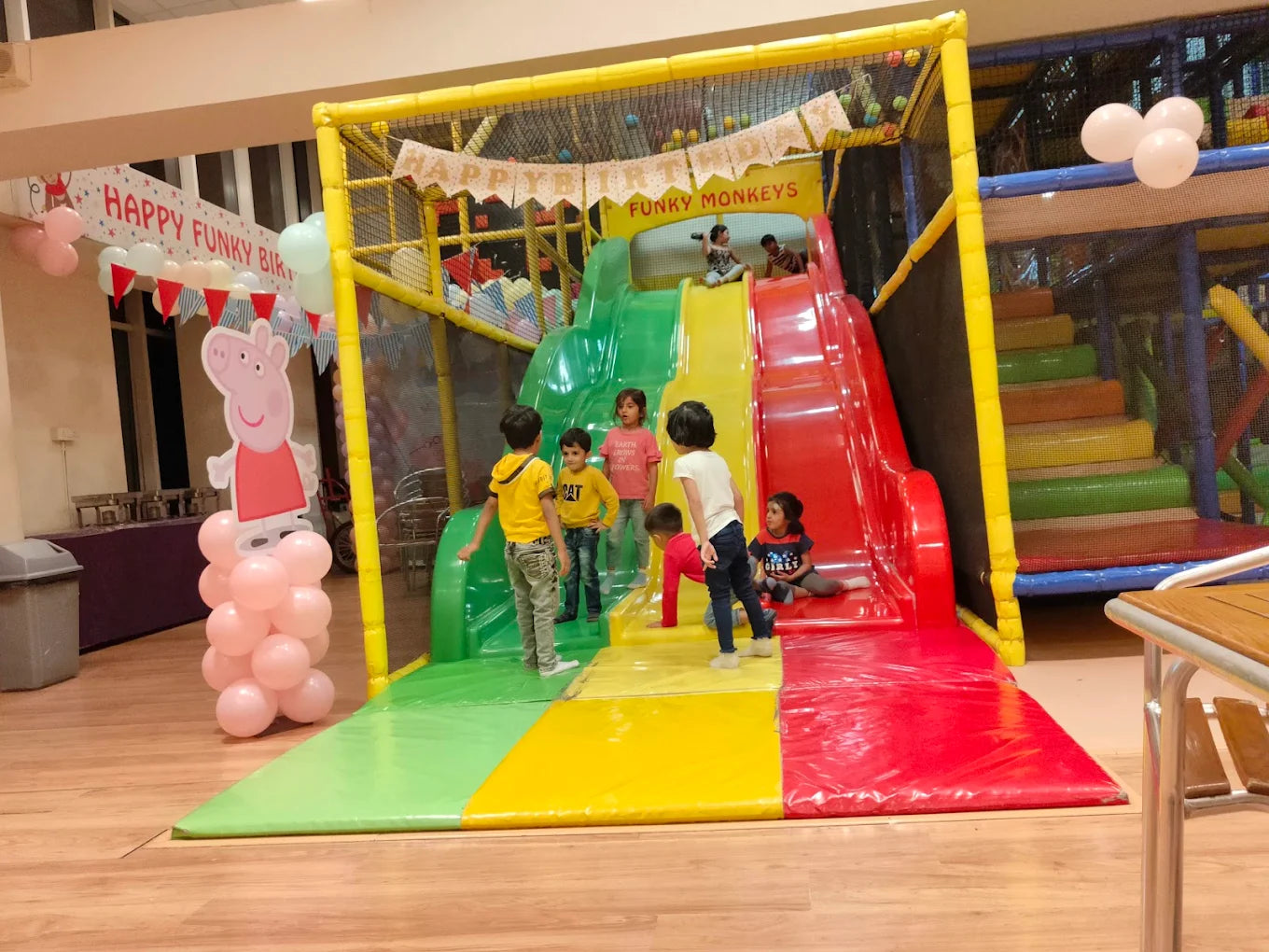 Children play on a colorful indoor playground slide. A Peppa Pig decoration stands nearby.