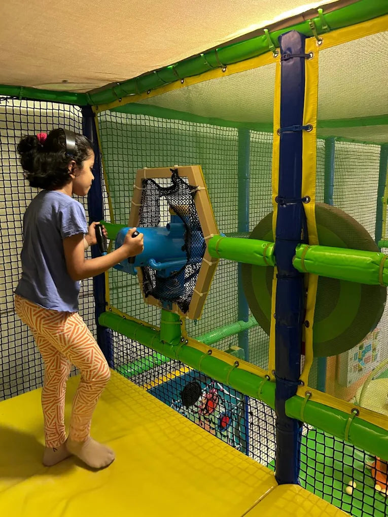 A young girl plays with a blue toy gun in a colorful indoor playground.