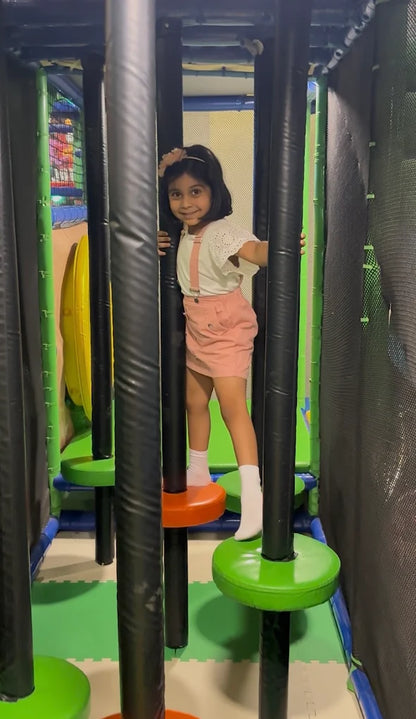A young girl smiles as she navigates a climbing structure with colorful platforms. She is holding onto black poles in a soft play area.