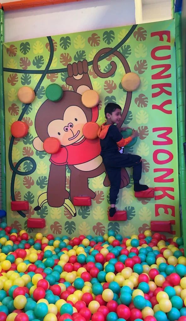 A young boy climbs a wall with monkey graphics, above a ball pit.