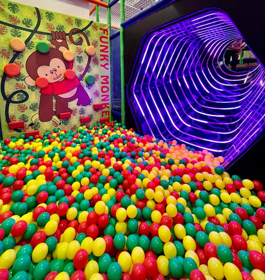A ball pit filled with colorful balls, next to a climbing wall with a monkey graphic and a purple-lit tunnel.