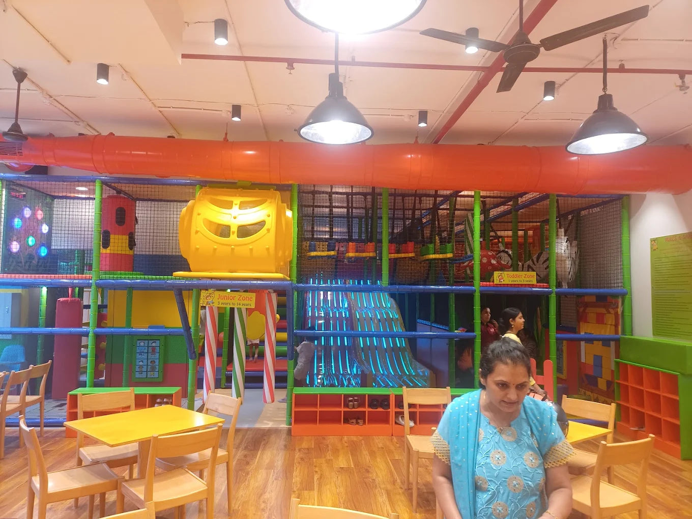 A woman sits at a table in a brightly colored indoor playground with a large play structure.