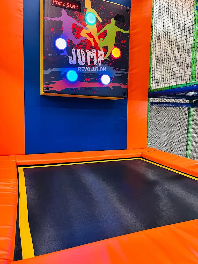 A trampoline park with a "Jump Revolution" game board and padded orange walls.