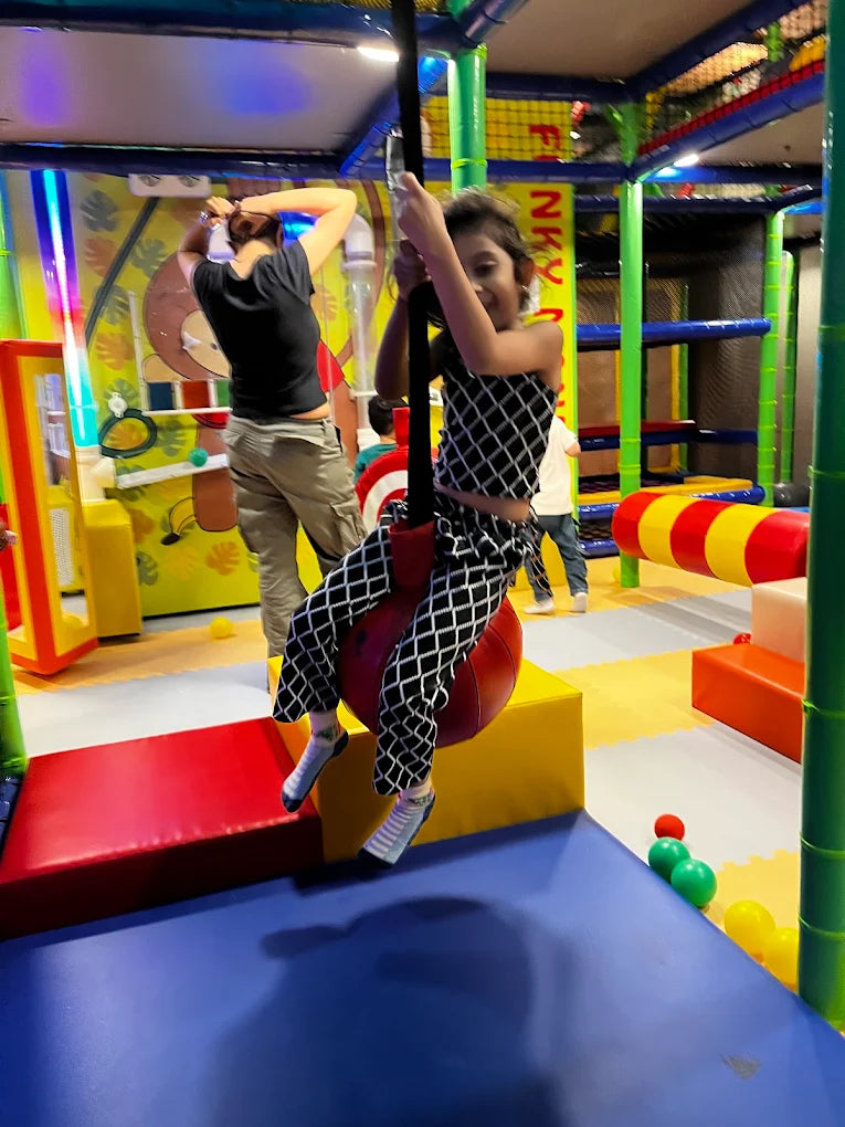 A young girl swings on a red ball in a colorful indoor playground while a woman stands nearby.