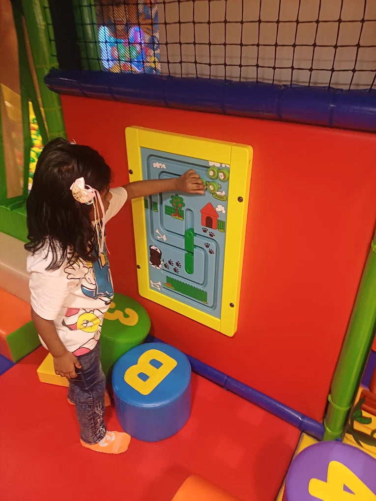 A young child plays with a colorful interactive wall panel in a soft play area. The panel features a maze with animal paw prints and a small red house.