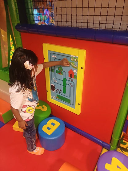 A young child plays with a colorful interactive wall panel in a soft play area. The panel features a maze with animal paw prints and a small red house.