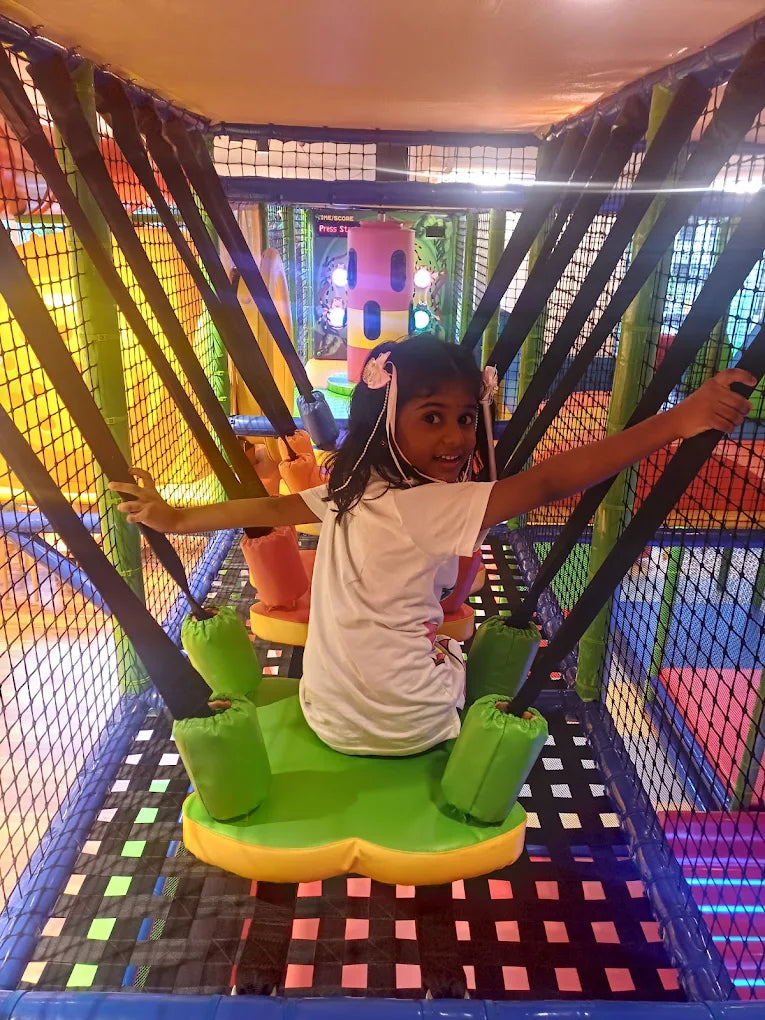 A young girl smiles while sitting on a colorful swing in an indoor playground.