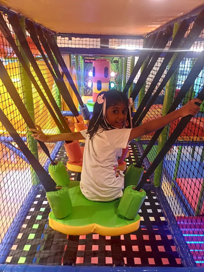 A young girl smiles while sitting on a colorful swing in an indoor playground.