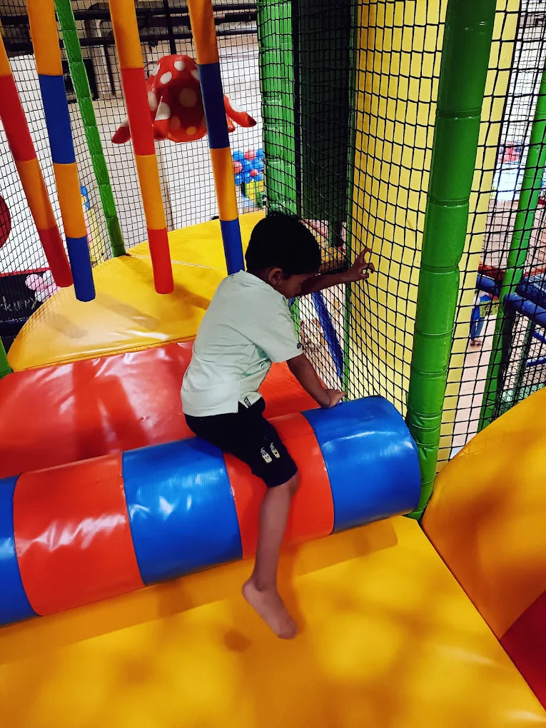A young boy plays in a colorful indoor playground, climbing on a soft cylindrical obstacle.