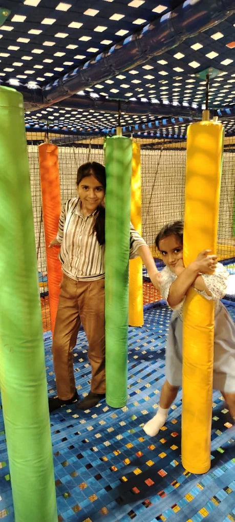 Two young girls play in a colorful indoor playground with large padded pillars.