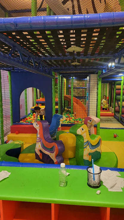 Children play in a colorful indoor playground with ball pits and soft play structures. A table in the foreground holds a water bottle and papers.