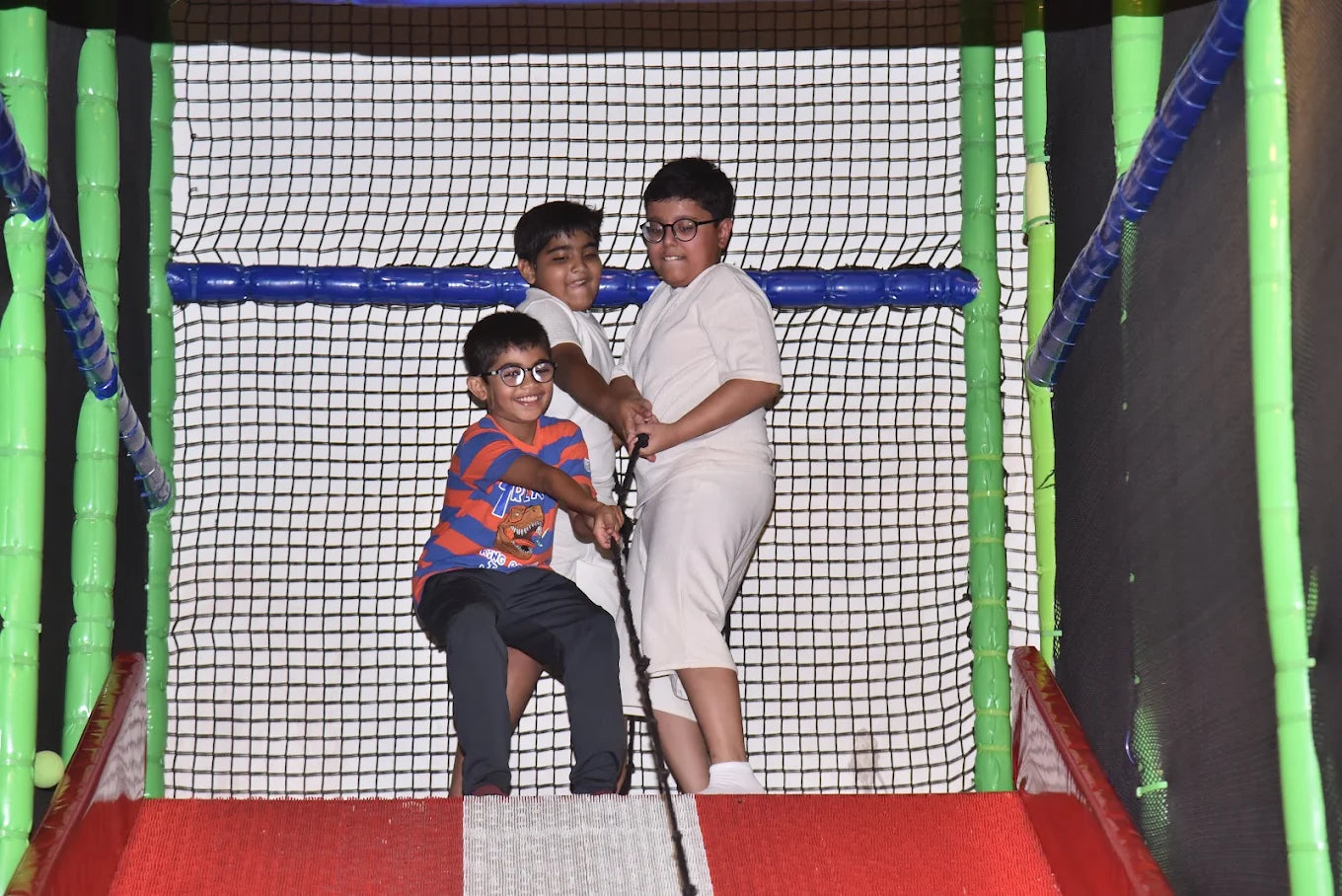 Three young boys play in a colorful indoor playground, pulling on a rope.