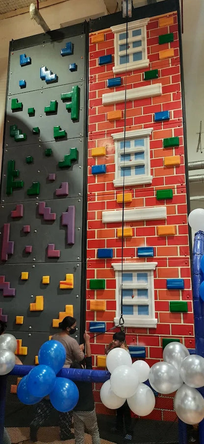 Children and adults climb a colorful indoor rock wall decorated like a brick building with windows. Balloons in blue and white are visible at the bottom.