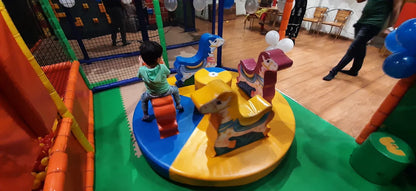 A young child rides a carousel in a colorful indoor playground. Other children's toys and balloons are visible.