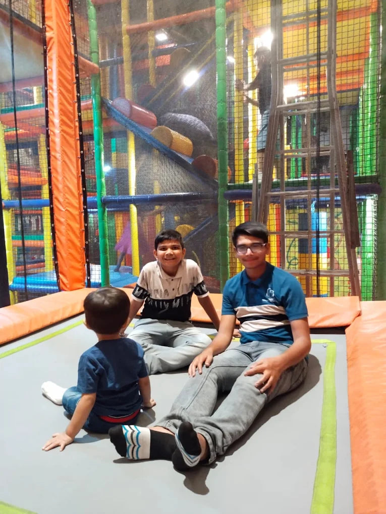 Three young boys sit on a trampoline in a colorful indoor play area.
