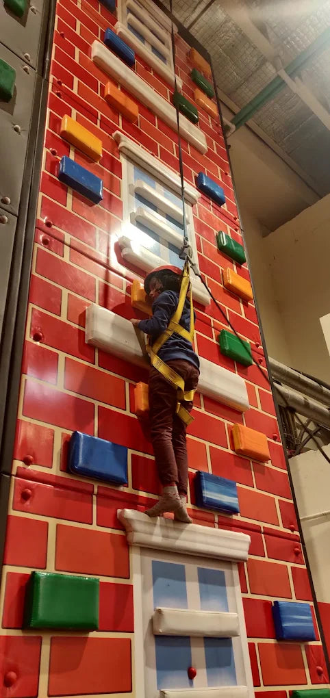 A child in a harness climbs a colorful indoor climbing wall designed to look like a brick building.