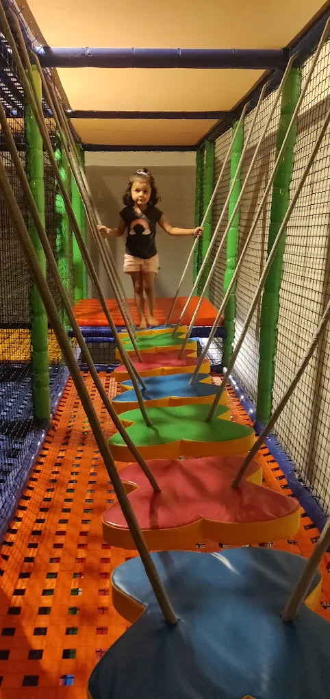 A young child walks across a colorful rope bridge in an indoor playground.