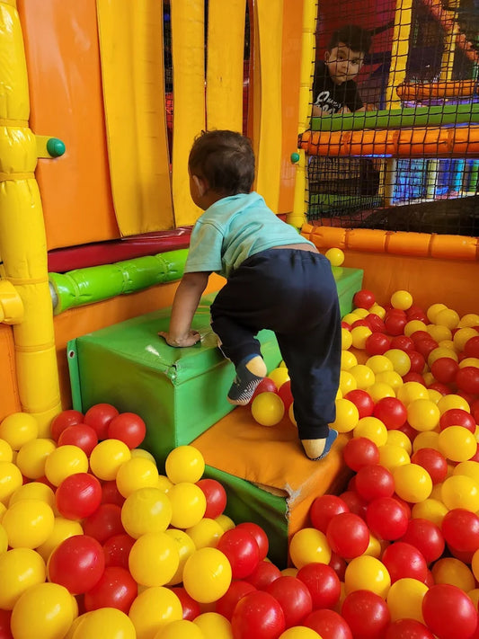 A young child climbs in a ball pit at an indoor playground, with another child visible in the background.