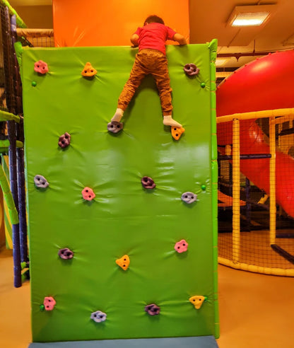 A child climbs a green bouldering wall in an indoor play area.